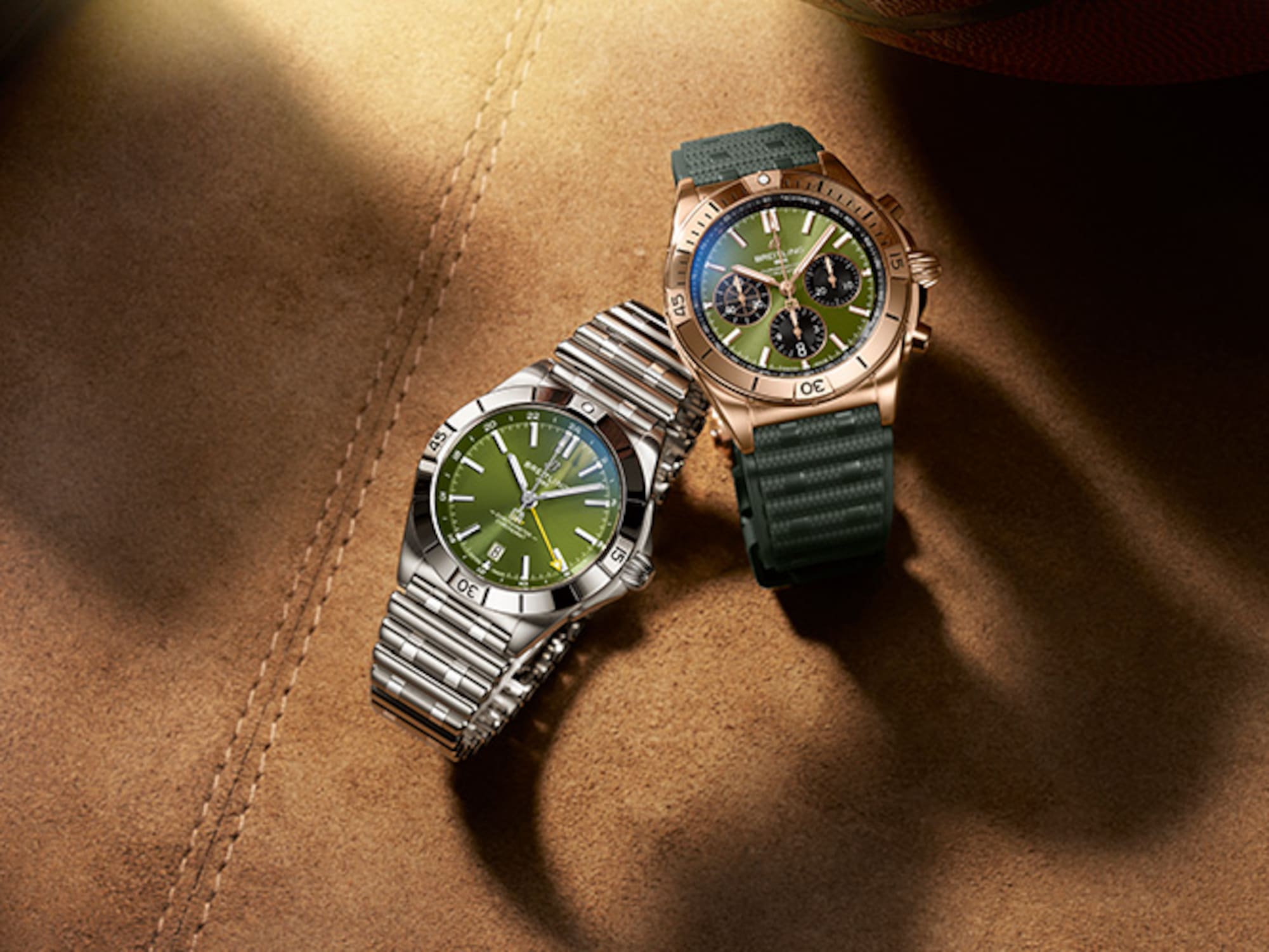 A gold and a silver Chronomat with dark green rubber straps on a brown table.