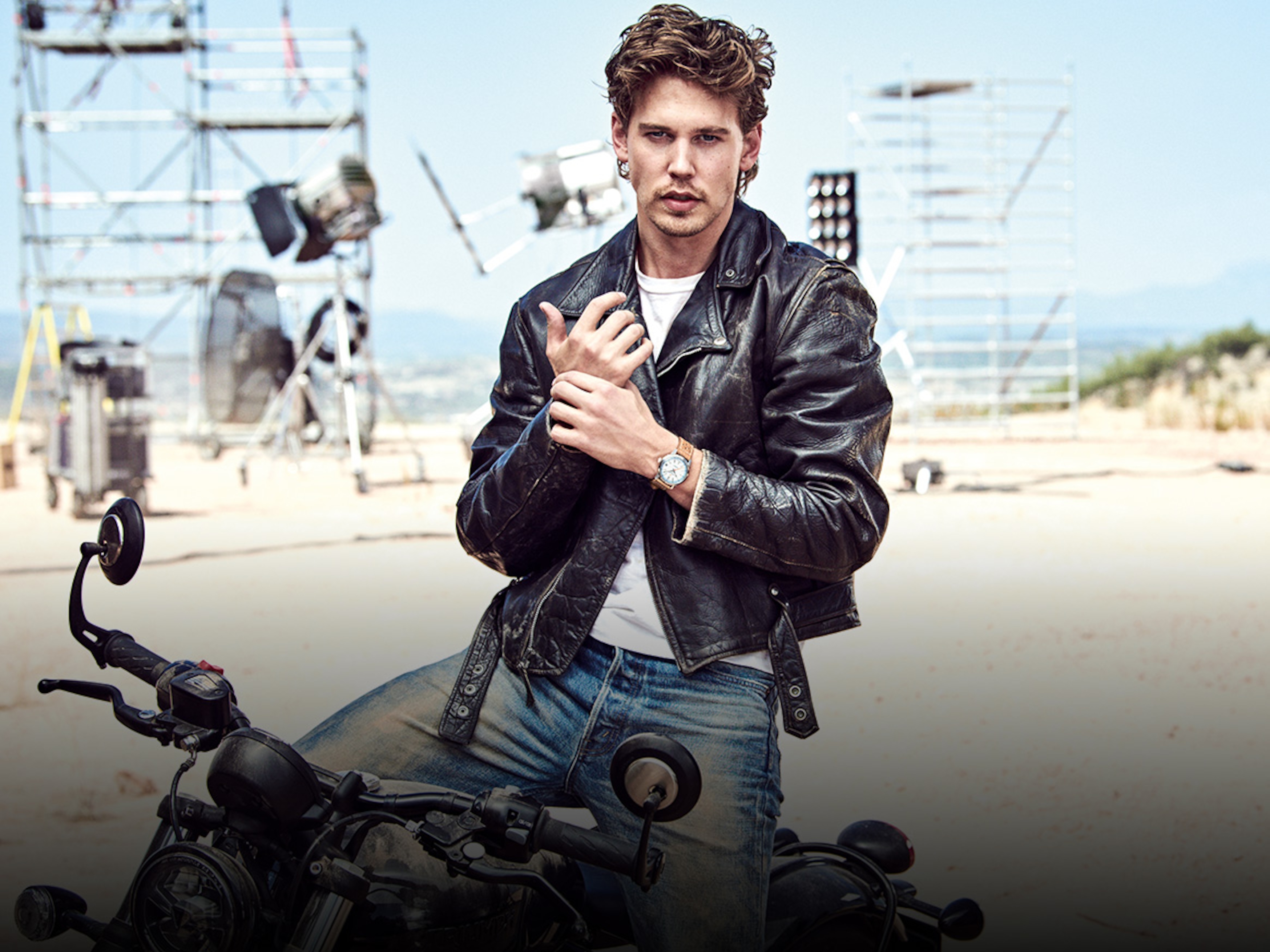 Person in black leather jacket wearing a Breitling watch, standing next to motorcycle on outdoor film set with lighting equipment.